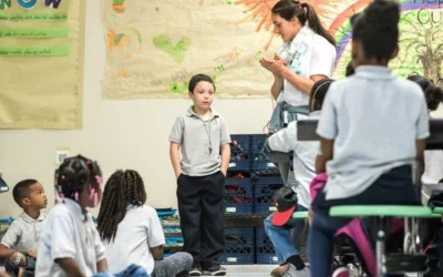 Photo of teacher applauding student standing in front of classroom with other students seated