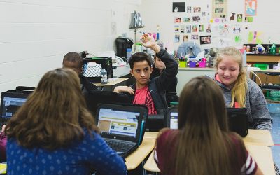 Student raises his hand, looking forward, while sitting between other students