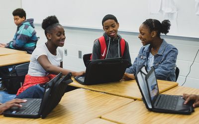 Three students, smiling, work together off of one laptop