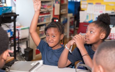 Two female students sit at desk in classroom as one raises her hand