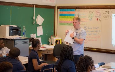 Teacher stands in front of classroom, pointing to paper in his hand as students look on