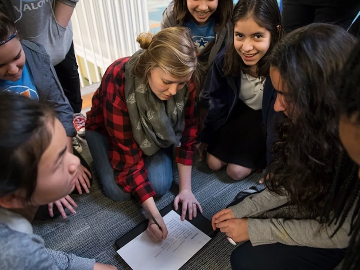 As teacher sits on the floor and writes on paper, students circle around her, looking down, while one student smiles at the camera