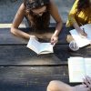 Three adults sit at outdoor table, reading books and taking notes